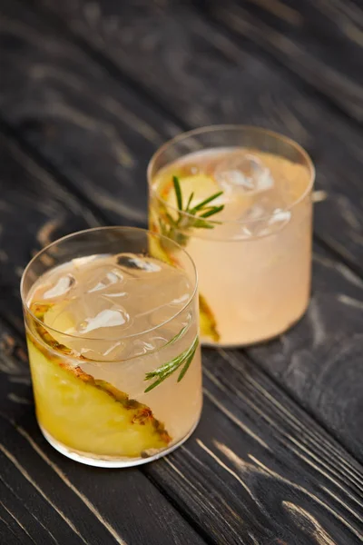 Due bicchieri di limonata con pezzetti di ananas, cubetti di ghiaccio e rosmarino su tavolo grigio in legno — Foto stock