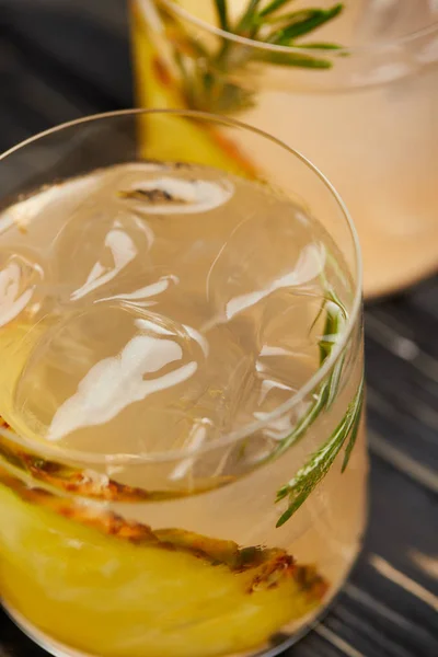 Enfoque selectivo de dos vasos de limonada con trozos de piña, cubitos de hielo y romero sobre mesa de madera gris - foto de stock