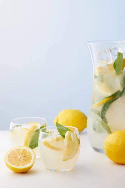 Jarra y dos vasos de limonada con hojas de menta, cubitos de hielo y rodajas de limón sobre fondo azul - foto de stock