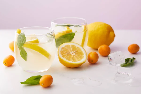 Dos vasos de limonada con hojas de menta, cubitos de hielo y rodajas de limón rodeados de kumquats y limones sobre fondo púrpura - foto de stock