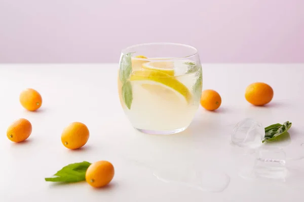 Glass of lemonade with mint leaves and lemon slices surrounded by ice cubes, kumquats on purple background — Stock Photo
