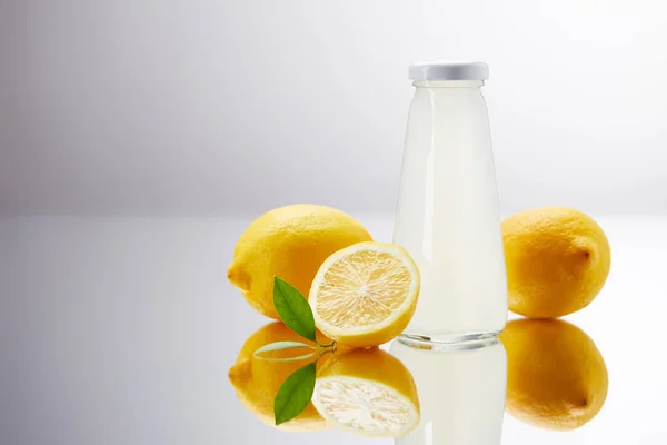 Glass bottle of delicious lemonade with lemons on reflective surface and on grey — Stock Photo