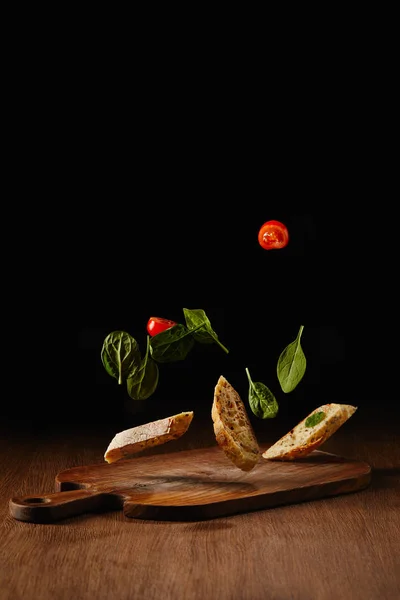 Trozos de pan y hojas de ensalada con tomates volando sobre la superficie de la mesa de madera - foto de stock