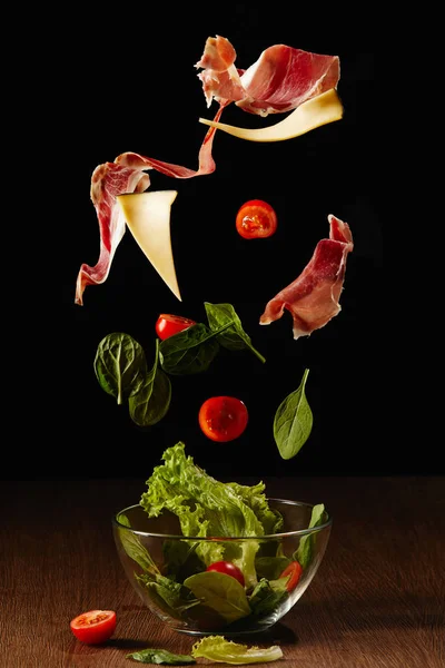 Ingredientes para ensalada de verduras y jamón cayendo en tazón sobre mesa de madera - foto de stock