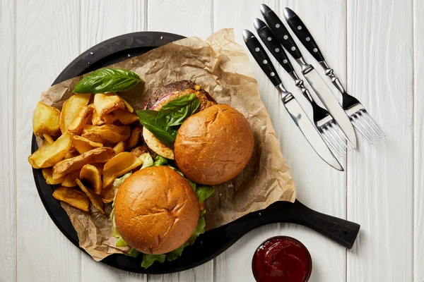 Blick von oben auf verführerische Hamburger und Pommes mit Sauce und Besteck auf weißem Tisch — Stockfoto