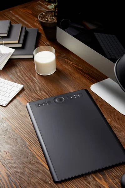 Vista de cerca del lugar de trabajo de diseñador gráfico con tableta gráfica, vaso de leche y cuadernos en la mesa de madera - foto de stock