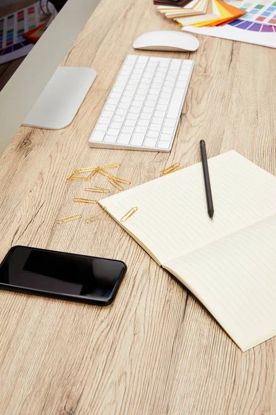 Vista de cerca del espacio de trabajo del diseñador con teléfono inteligente, portátil vacío, pantalla del ordenador y teclado en la superficie de madera - foto de stock