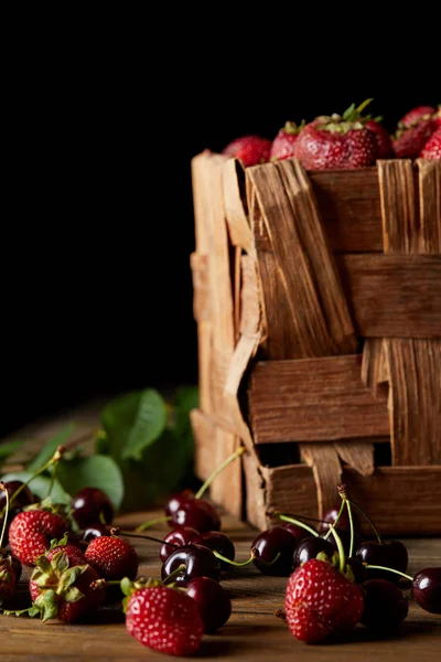 Спелые вишни и клубника на деревянной поверхности с листьями и деревенская коробка на черном — стоковое фото