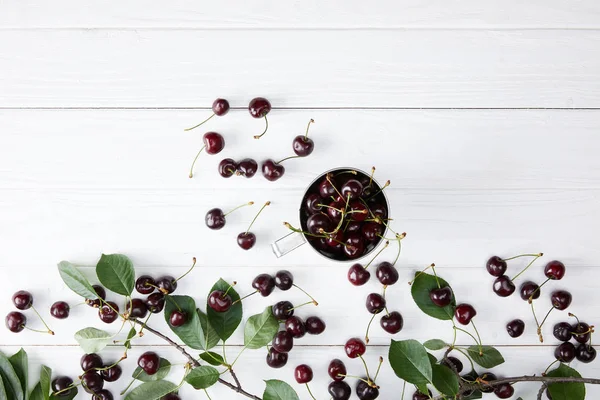 Draufsicht auf frische reife Kirschen in Tasse auf weißer Holztischplatte mit Blätterzweig — Stockfoto