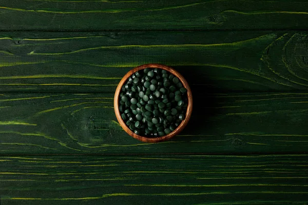 Vue du dessus du bol en bois avec pile de pilules de spiruline sur la table en bois vert — Photo de stock