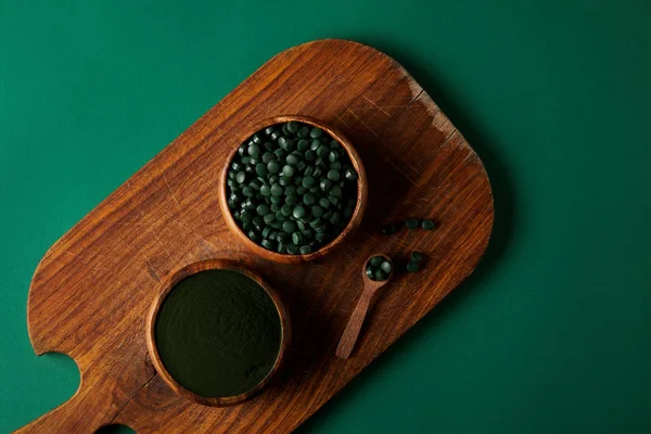 Top view of cutting board, wooden spoon,  bowls with spirulina powder and spirulina in pills on green table — Stock Photo