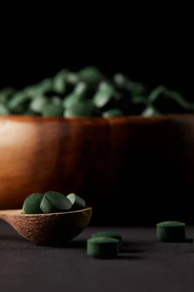 Enfoque selectivo de cuchara de madera y tazón con pila de píldoras de espirulina sobre fondo negro - foto de stock