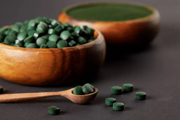 Selective focus of wooden spoon and bowls with ground spirulina and spirulina in pills on grey table — Stock Photo