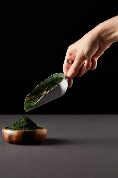 Cropped image of woman throwing spirulina powder by scoop into wooden bowl on black background — Stock Photo