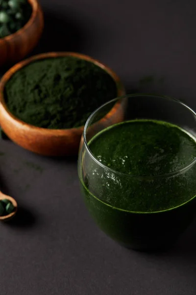 Selective focus of wooden spoon, glass of fresh smoothie from spirulina, bowls with spirulina powder and spirulina pills on grey table — Stock Photo