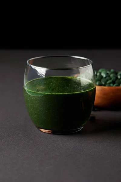 Glass of fresh smoothie from spirulina and wooden bowl with spirulina pills on black background — Stock Photo