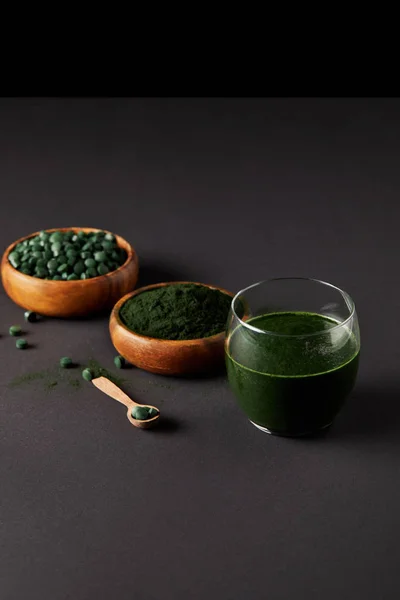 Cuillère en bois, verre de smoothie à la spiruline fraîche, bols avec poudre de spiruline et pilules de spiruline sur table grise — Photo de stock