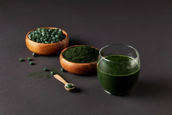 Arranged wooden spoon, glass of fresh smoothie from spirulina, bowls with spirulina powder and spirulina pills on grey table — Stock Photo