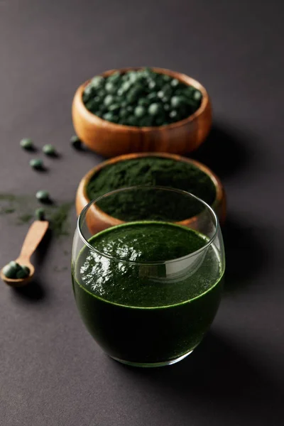 Selective focus of arranged wooden spoon, glass of spirulina smoothie, bowls with spirulina powder and spirulina pills on grey table — Stock Photo