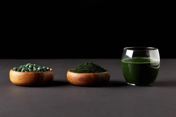 Glass of spirulina smoothie, bowls with spirulina powder and spirulina pills on black background — Stock Photo
