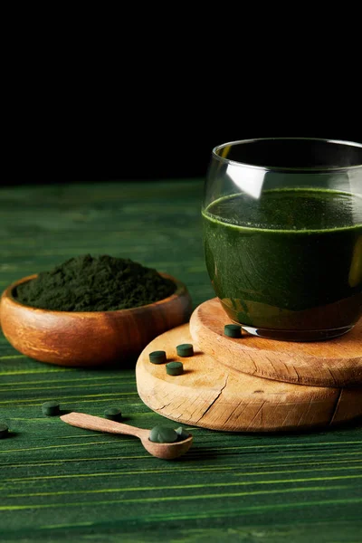 Wooden slices with fresh spirulina drink in glass, wooden spoon and spirulina powder in wooden bowl on black background on green wooden table — Stock Photo