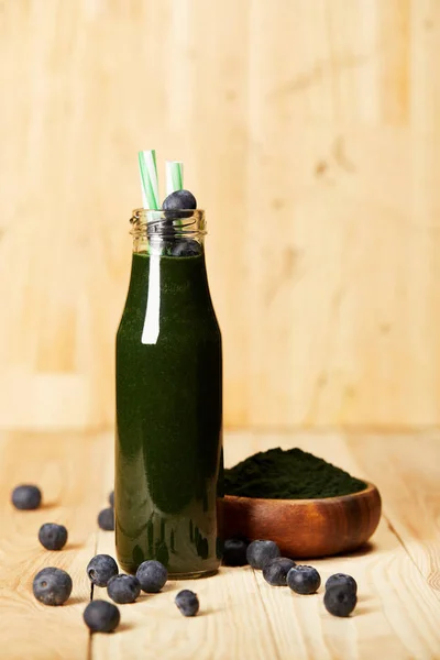 Schüssel mit Spiruluna-Pulver, Flasche Spirulina-Smoothie mit Blaubeeren und Trinkhalm auf Holztisch — Stockfoto