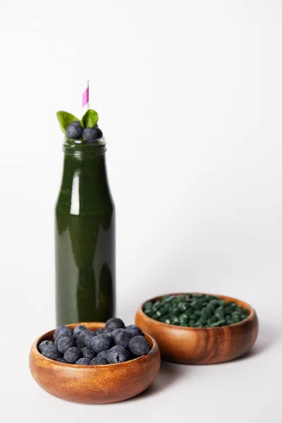 Selektiver Fokus einer Flasche Spirulina-Smoothie mit Minzblättern und Trinkhalm, Schalen mit Blaubeeren und Spirulina-Pillen auf grauem Hintergrund — Stockfoto