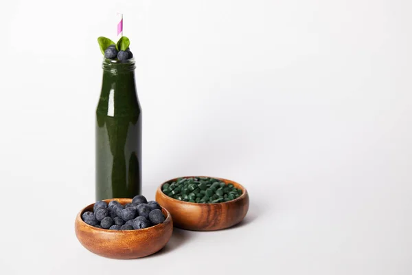 Bottle of spirulina smoothie with mint leaves and drinking straw, bowls with blueberries and spirulina pills on grey background — Stock Photo