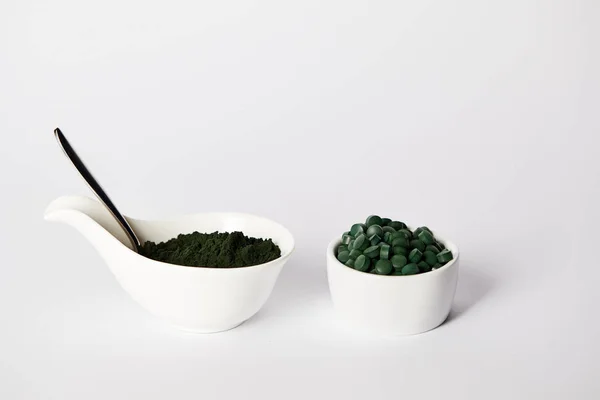 Cuillère, poudre de spiruline et pilules de spiruline dans des bols sur fond gris — Photo de stock