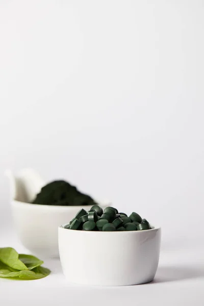 Selective focus of leaves, bowls with spirulina pills and spirulina powder on grey background — Stock Photo