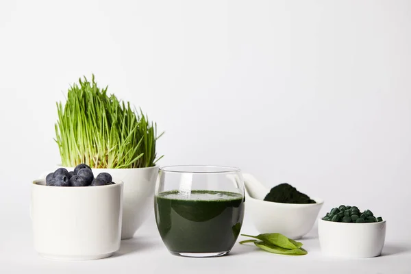 Fresh smoothie from spirulina, cups with spirulina grass and blueberries, leaves, bowls with spirulina powder and spirulina pills on grey background — Stock Photo