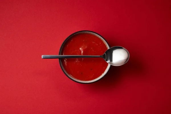 Vue du dessus de la délicieuse soupe de tomates rouges dans une assiette et une cuillère sur la table rouge — Photo de stock