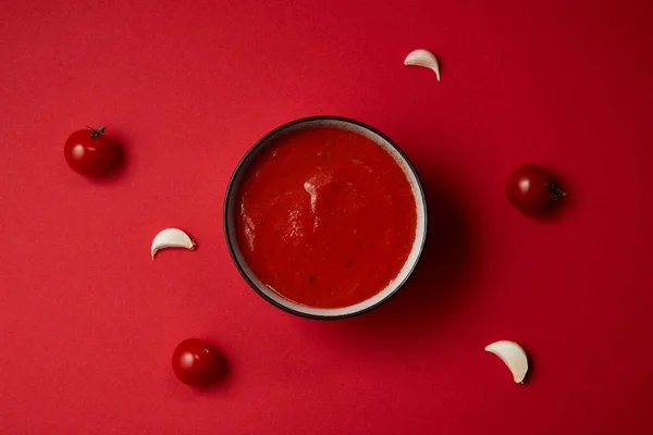 Vue du dessus de la soupe de tomates savoureuse dans une assiette et l'ail et les tomates éparpillées sur la table rouge — Photo de stock