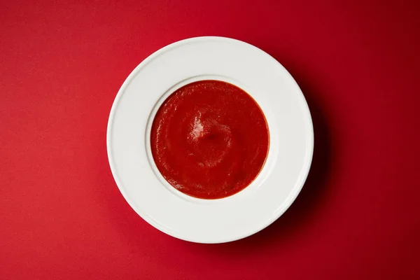 Vue du dessus de la soupe de tomates savoureuse dans une assiette blanche sur la table rouge — Photo de stock