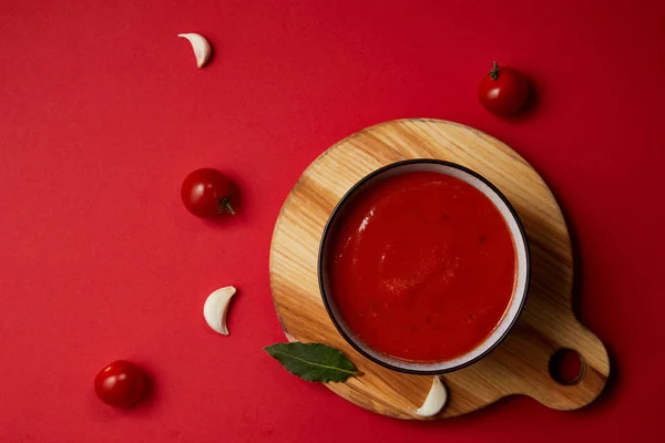 Вид сверху на вкусный томатный суп в тарелке на разделочной доске на красном столе — стоковое фото