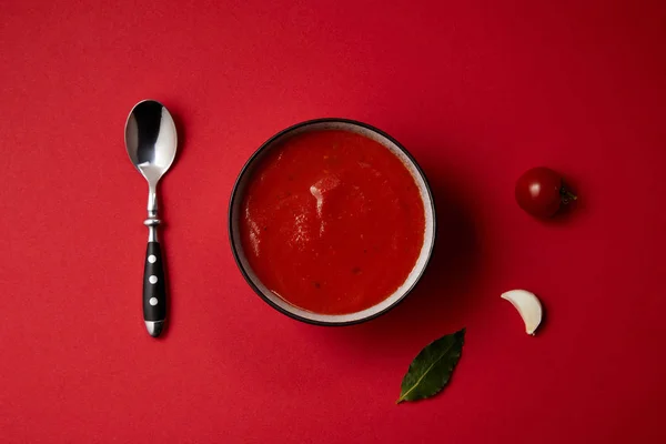 Вид зверху смачний томатний суп в тарілці, ложці, свіжий помідор і часник на червоному столі — стокове фото