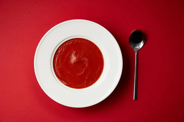 Elevated view of tomato soup in plate and spoon on red table — Stock Photo