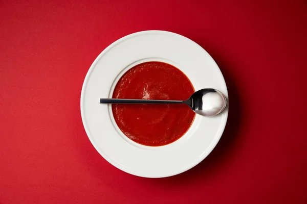 Vista superior da sopa de tomate na placa e colher na mesa vermelha — Fotografia de Stock