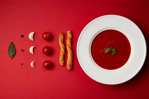 Flat lay of tomato soup in plate and spices, vegetables with bread sticks on red table — Stock Photo