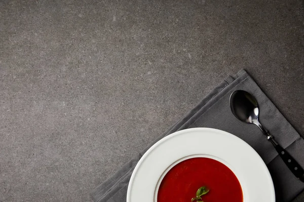 Vista superior de la sabrosa sopa de tomate en plato y cuchara en servilleta gris sobre mesa gris - foto de stock