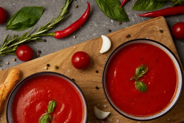 Вид на вкусный томатный суп в тарелках на разделочной доске с овощами на сером столе — стоковое фото