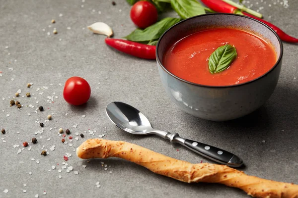 Cooked tomato soup and chili peppers, basil and bread stick on grey table — Stock Photo