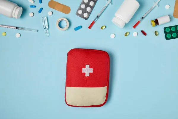 Vista superior de la bolsa roja del botiquín de primeros auxilios en la superficie azul con diferentes medicamentos - foto de stock