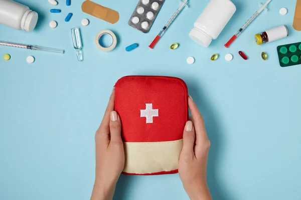 Tiro recortado de la mujer que sostiene la bolsa del kit de primeros auxilios sobre la superficie azul con varios medicamentos - foto de stock