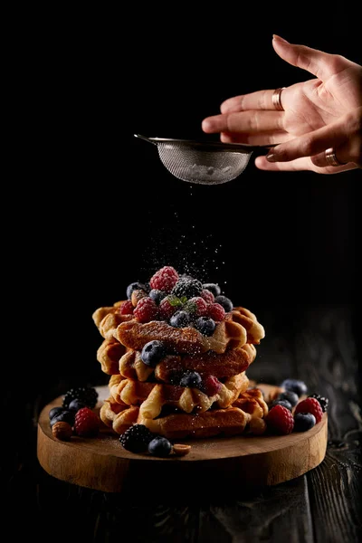 Cropped shot of woman spilling sugar powder onto belgian waffles through sieve on black — Stock Photo