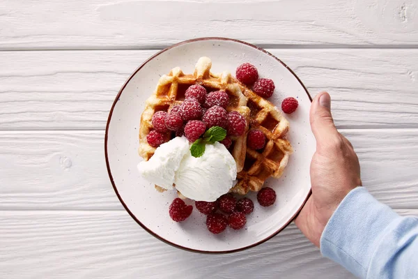 Plan recadré d'un homme tenant une assiette de gaufres belges avec des framboises et de la crème glacée sur une table en bois blanc — Photo de stock