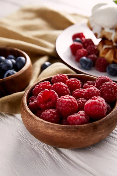 Close-up shot of bowls with berries with belgian waffles blurred on background on white wooden table — Stock Photo