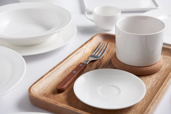 Selective focus of wooden tray, fork with various plates, bowl and cup on white table — Stock Photo