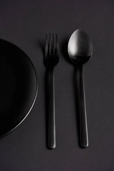 Вид зверху чорна ложка, виделка і тарілка на чорному столі, мінімалістична концепція — стокове фото