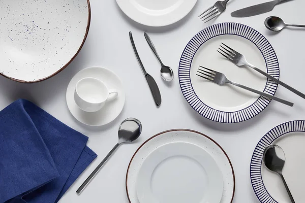 Вид сверху на различные керамические плиты, чашки, кухонное полотенце, вилки, ложки и нож на белый стол — стоковое фото